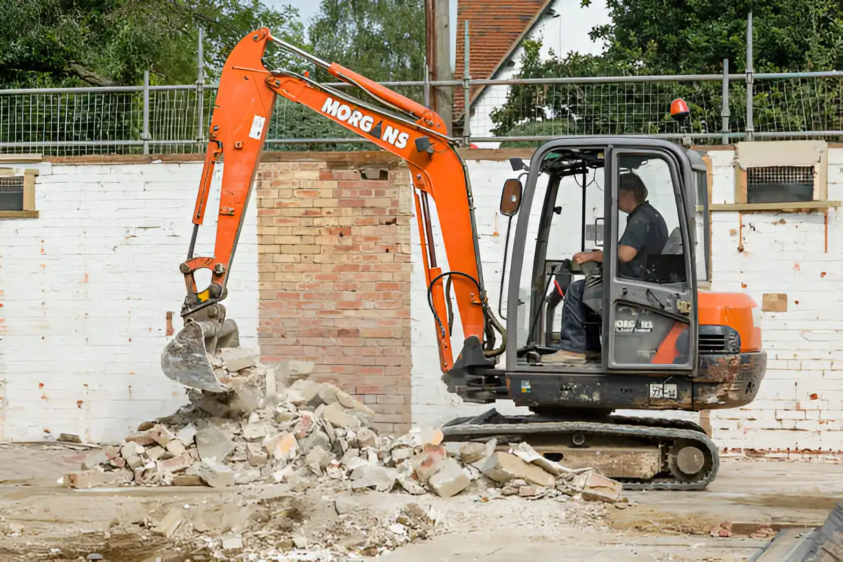 An orange excavator operated by a construction worker demolishing a brick wall, with rubble being cleared on a construction site.