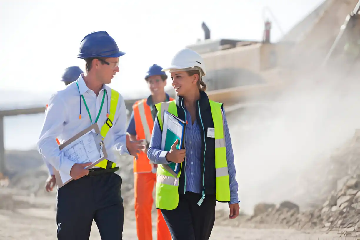 Project managers and engineers in safety gear discussing plans on a demolition site, with heavy machinery in the background.