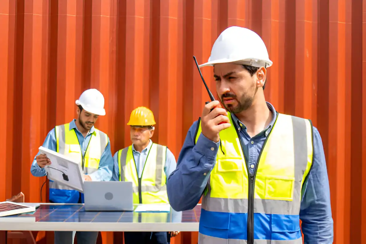 Group of construction workers wearing safety vests and hard hats, communicating and planning at a job site. One worker is using a walkie-talkie, while others review plans on a laptop, emphasizing teamwork and construction management.