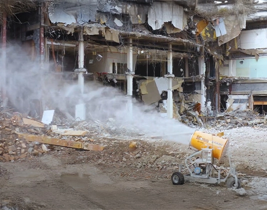 A demolition site showing extensive structural damage with debris scattered around. A dust suppression machine is actively spraying mist to control dust during the demolition process.