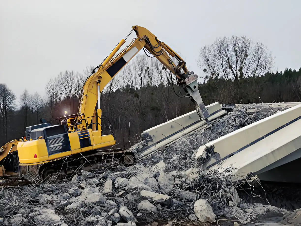 A yellow excavator actively demolishing a concrete structure, surrounded by rubble and debris, with trees in the background, illustrating heavy machinery in demolition work.