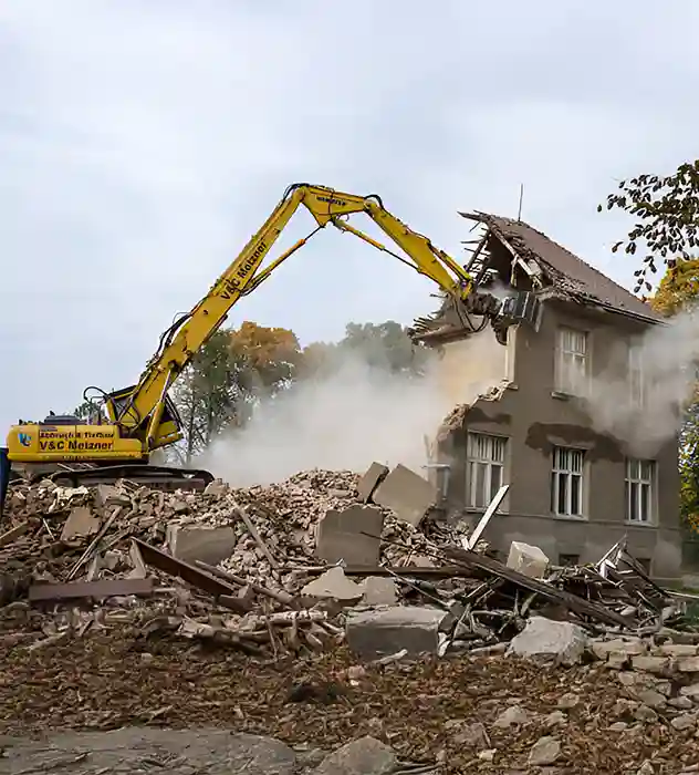 A large yellow excavator demolishing an old two-story building, with rubble and debris scattered around. Dust clouds form as the structure is being torn down, with trees visible in the background.