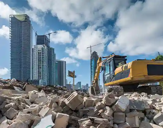 A construction site in an urban area with an excavator amidst rubble. Tall skyscrapers and cranes are visible in the background under a blue sky with clouds.