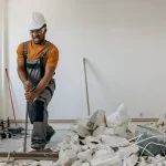 Construction worker in orange shirt and overalls using demolition bar to remove flooring during renovation