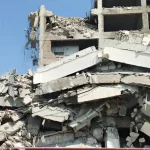 Partially collapsed concrete building with rubble and debris under a clear blue sky.