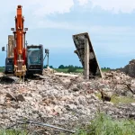 Orange excavator on demolition site with concrete rubble and debris against rural landscape background
