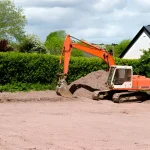 Orange excavator leveling soil near a residential area for site preparation.