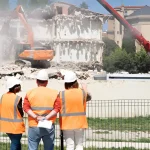 Workers in safety vests observe building demolition with excavator and crane