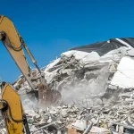 Excavator removing rubble from a demolished building under a clear blue sky.