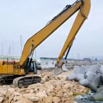 Yellow excavator clearing rocky coastal site, moving large concrete blocks near water