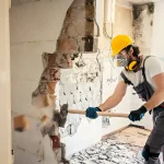 Worker in hard hat breaking brick wall with hammer, demolition safety equipment