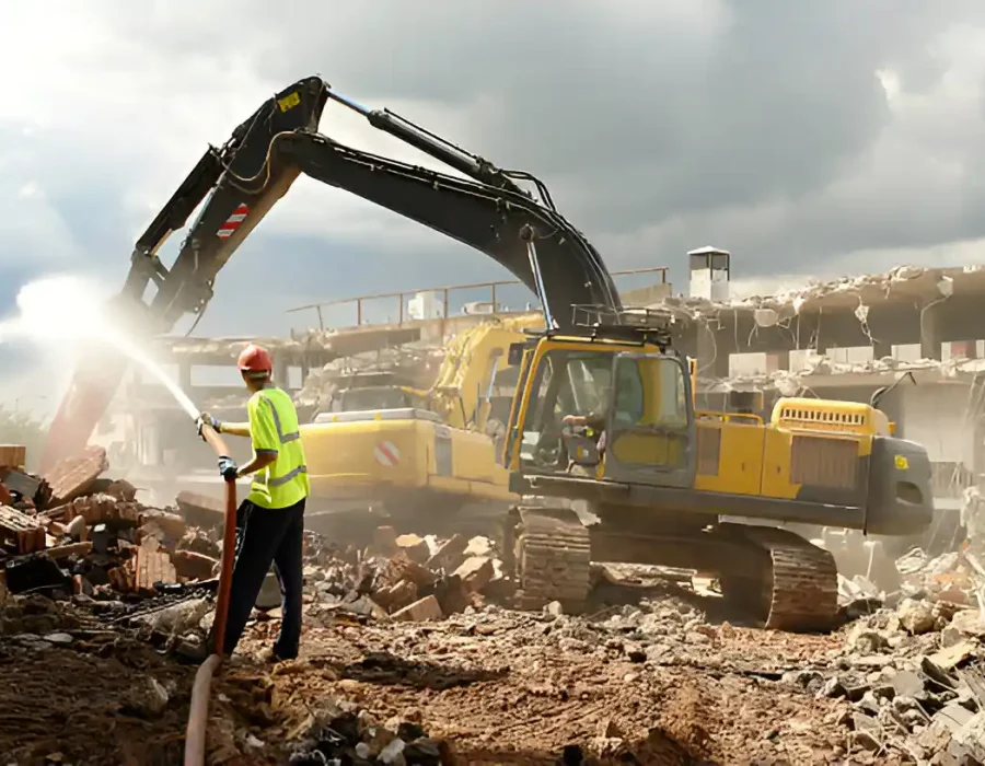 Safety worker supervising excavator demolition with dust control measures at commercial site