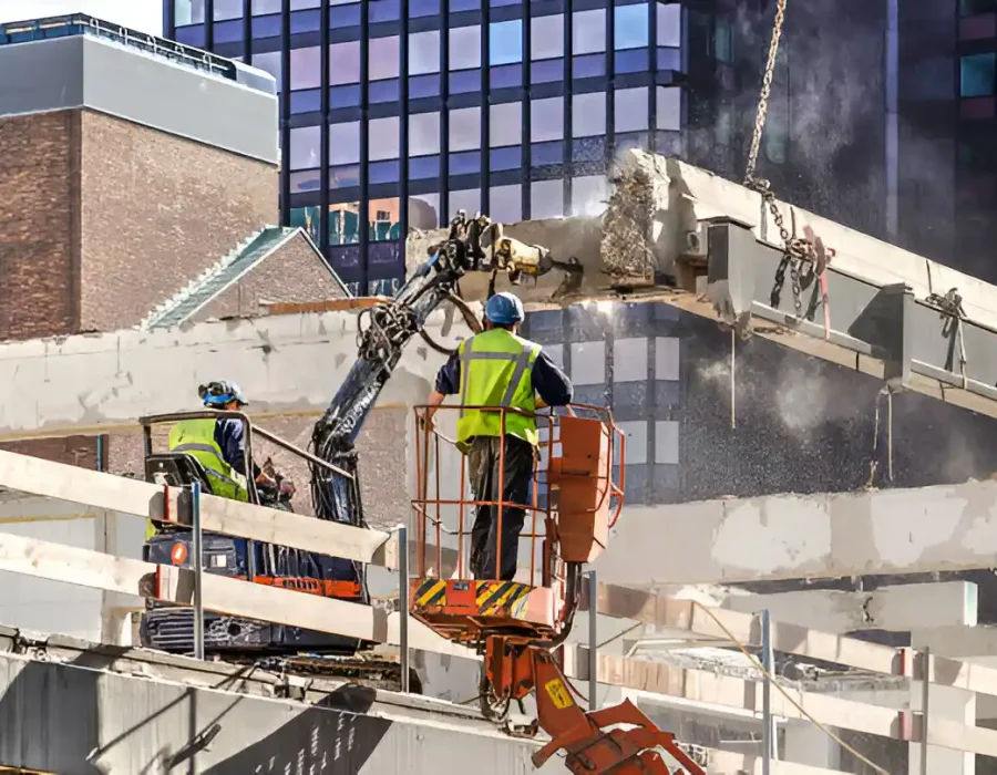 Workers in safety gear operating lift and machinery to demolish concrete structure near glass office building