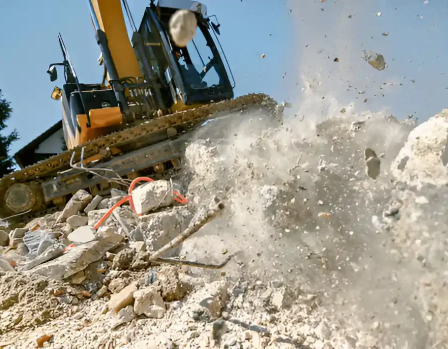 Yellow excavator breaking concrete wall with dramatic flying debris and dust against blue sky background