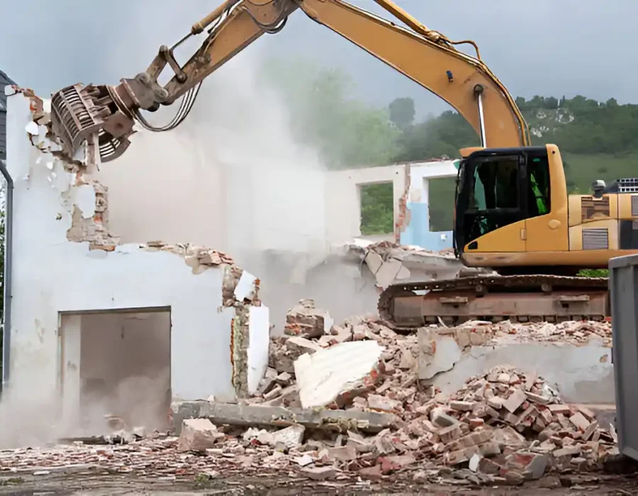 Yellow excavator demolishing white brick house, debris pile, dumpster in background