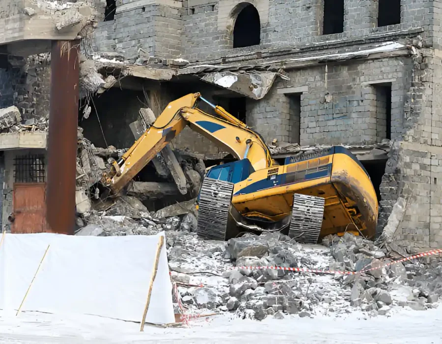 Yellow excavator dismantling stone building, debris field, safety barriers in foreground