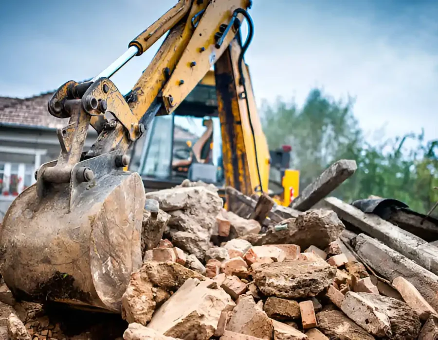 Yellow excavator bucket crushing concrete rubble near residential building