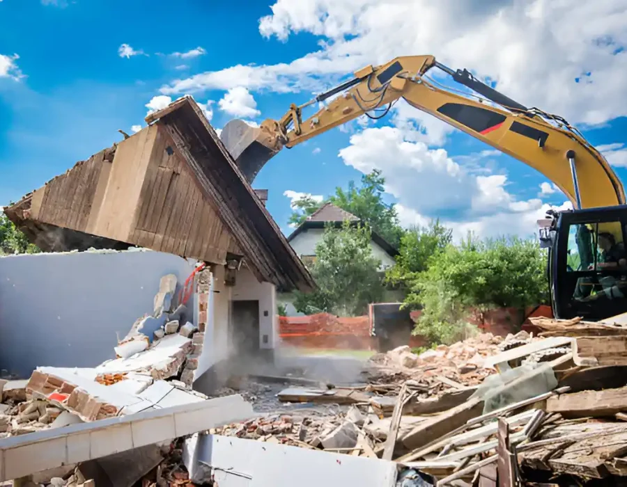Yellow excavator dismantling old wooden house with debris scattered around