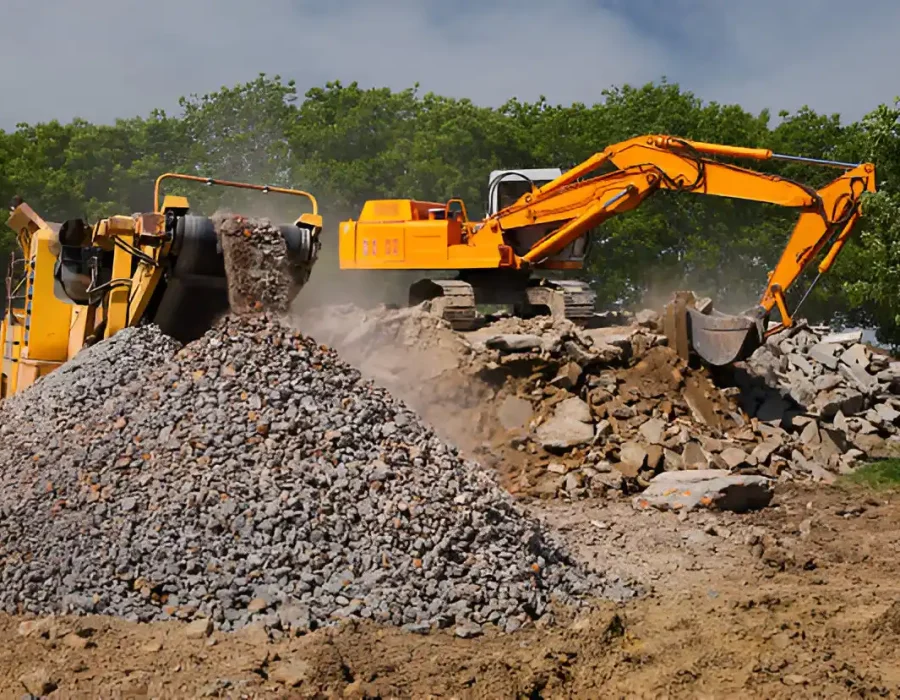 Excavator loading rubble into a crusher machine, creating a pile of processed debris for removal.