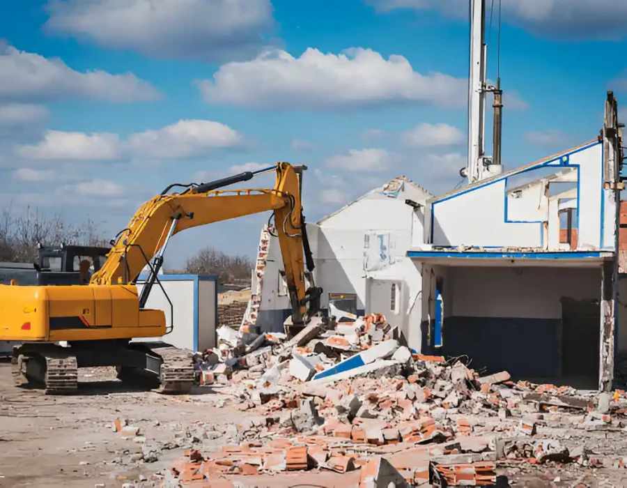 Yellow excavator demolishing white commercial building with brick debris pile on cleared site