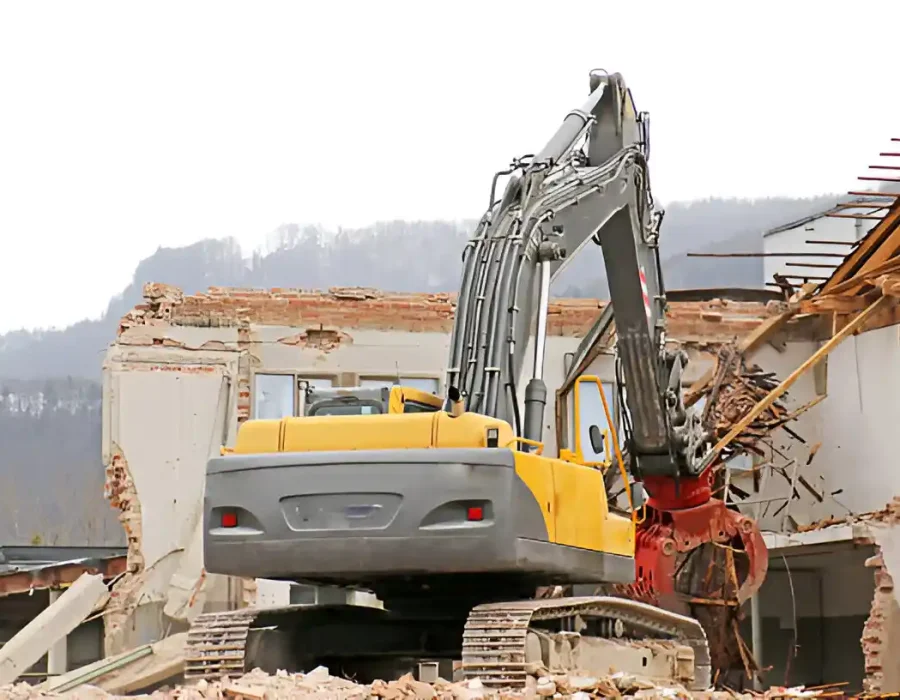 Yellow excavator with hydraulic grapple demolishing brick building in mountainous setting