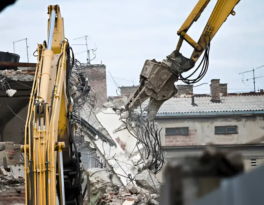 Two yellow excavator arms demolishing concrete building with exposed rebar and structural debris