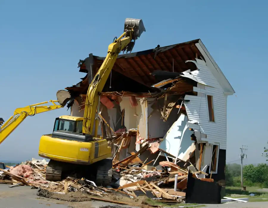 Yellow excavator tearing down white wooden house during complete residential demolition service