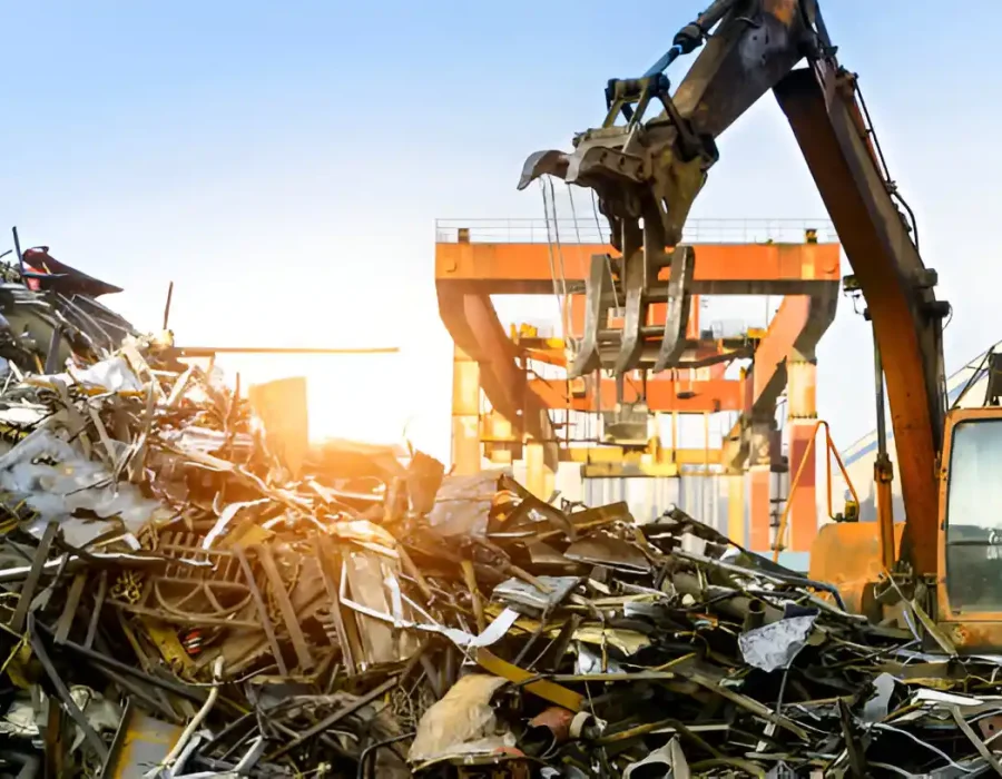 Excavator with grapple sorting metal debris at recycling yard during sunset with crane in background