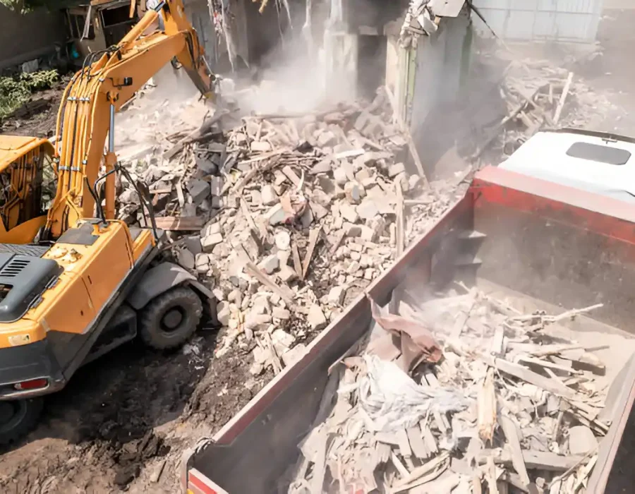 Excavator and dump truck working together to clear mixed demolition debris amid dust clouds