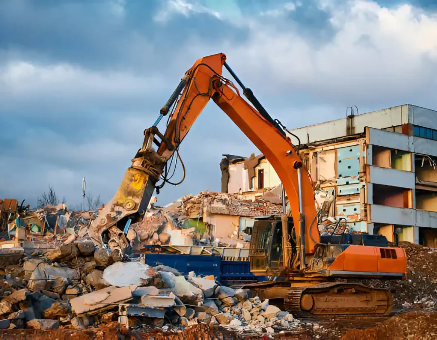 Orange excavator demolishing multi-story building with concrete rubble piles in foreground