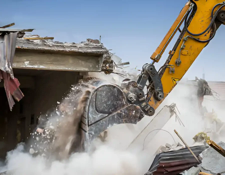 Yellow excavator demolishing building roof, debris and dust flying during demolition process