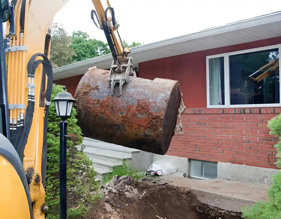 Yellow excavator lifting rusty oil tank from residential foundation with red brick house and landscaping visible