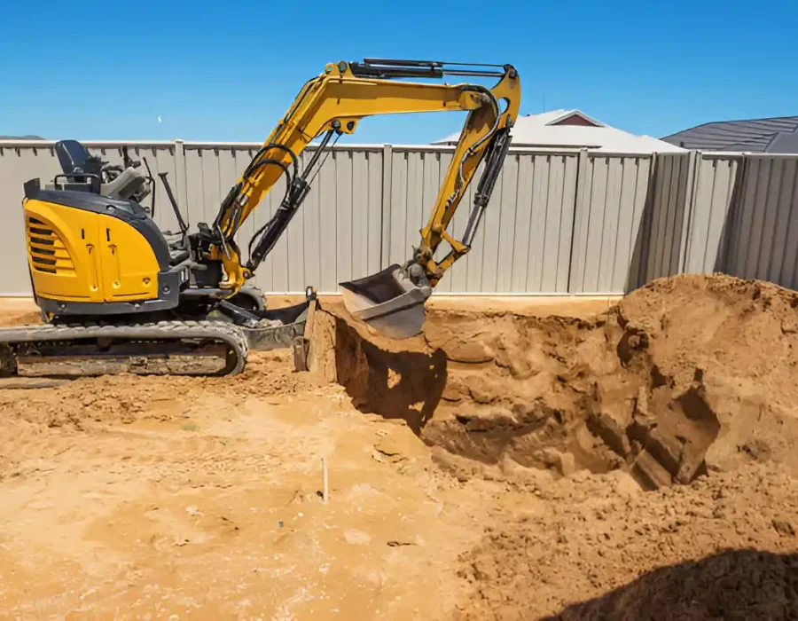 Yellow compact excavator digging sandy soil against grey fence backdrop for residential site preparation