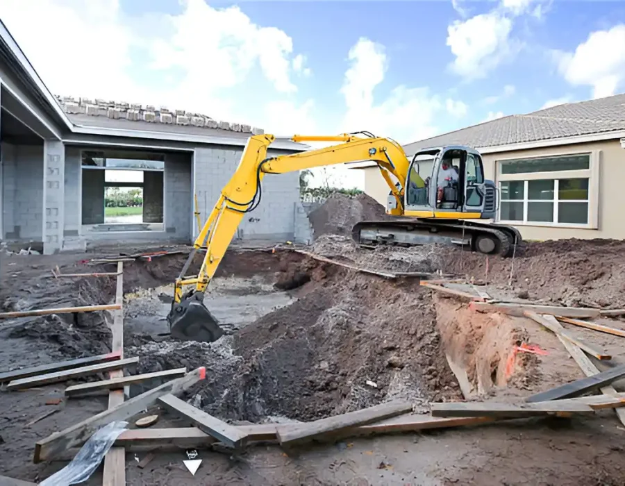 Yellow excavator digging foundation trench between concrete block building and beige structure with scattered debris