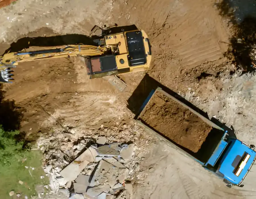 Aerial view of yellow excavator loading demolition debris into blue dump truck with trees along worksite