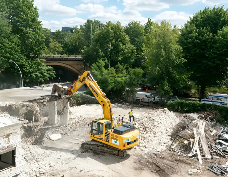Yellow excavator removing debris from building, site cleanup in urban area with trees in background