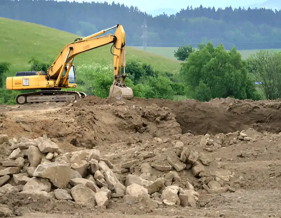 Yellow excavator on hillside clearing rocky terrain with mountain forest backdrop and stone debris piles