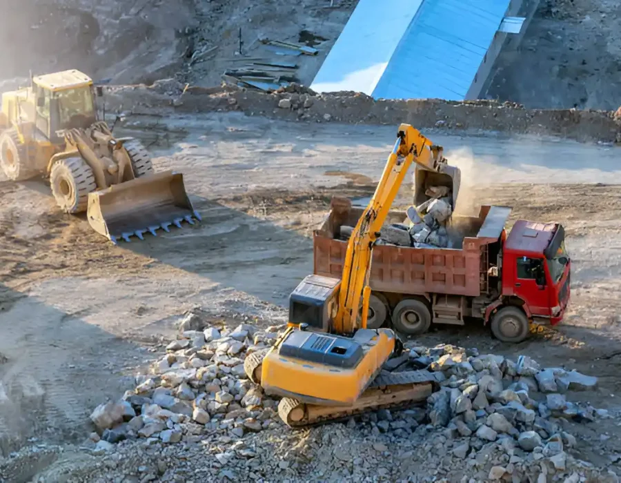Yellow excavator loading concrete debris into red dump truck with front loader and blue building in background