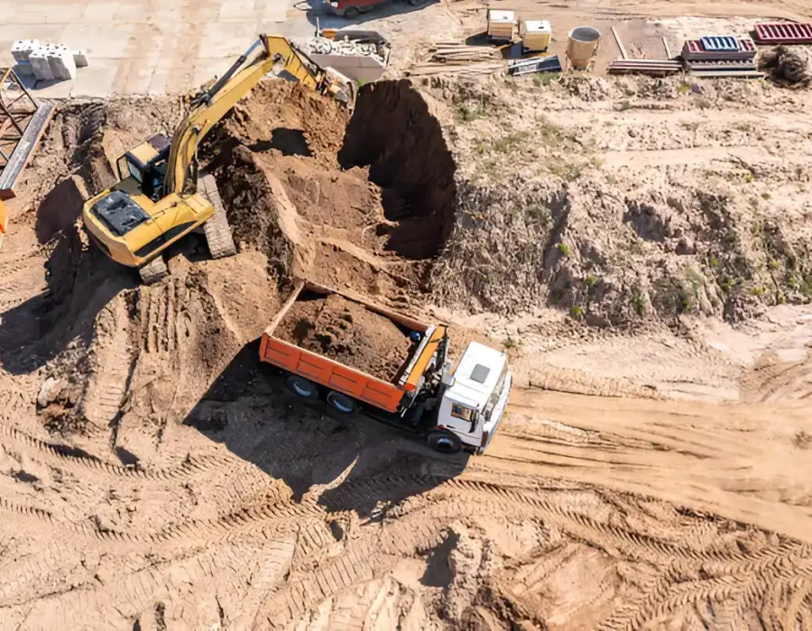 Yellow excavator loading dirt into white dump truck at sandy construction site, aerial view of earthwork progress