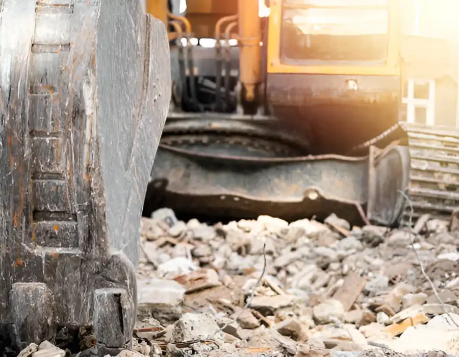 Close-up of excavator attachment amid scattered concrete and building debris with warm sunlight filtering in