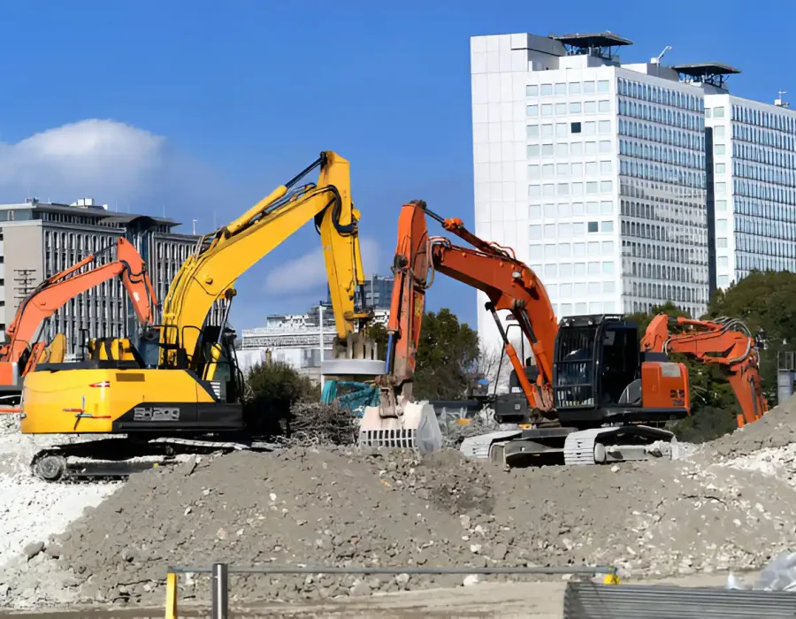 Yellow and orange excavators working on demolition site with modern office buildings in urban backdrop