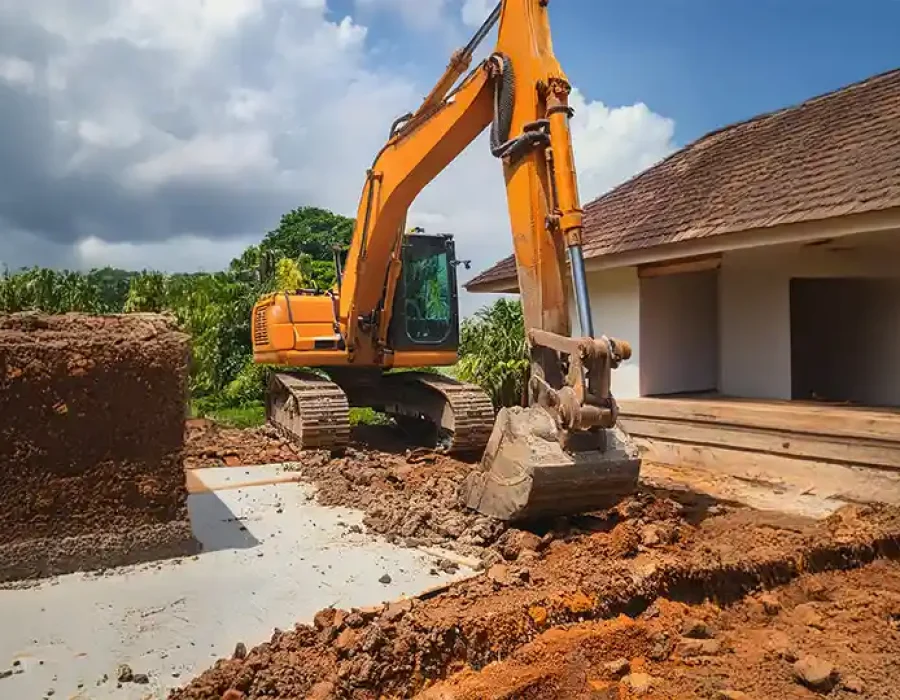 Excavator performing residential demolition and site preparation, removing concrete and soil near house foundation