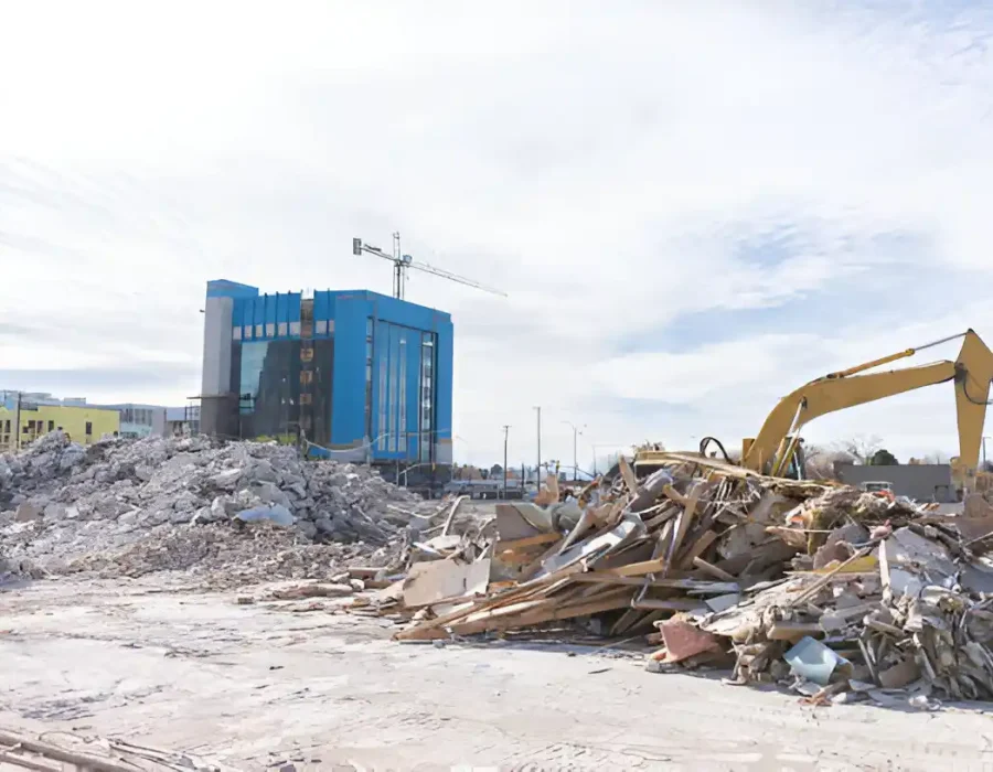 Yellow excavator sorting construction waste near blue building with debris piles