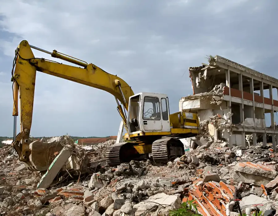 Yellow excavator dismantling two-story concrete building with massive debris pile