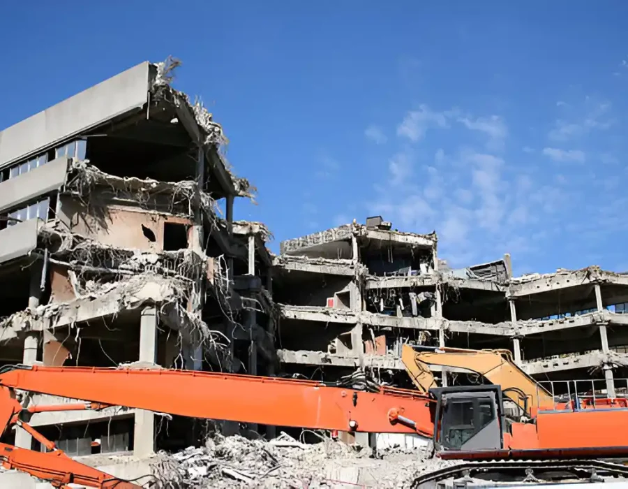 Orange long-reach excavator dismantling multi-story concrete building against blue sky