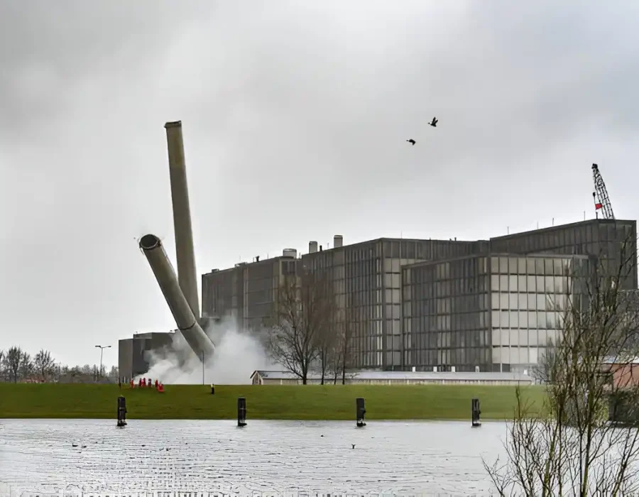 Precision building demolition with falling smoke stacks near modern industrial complex