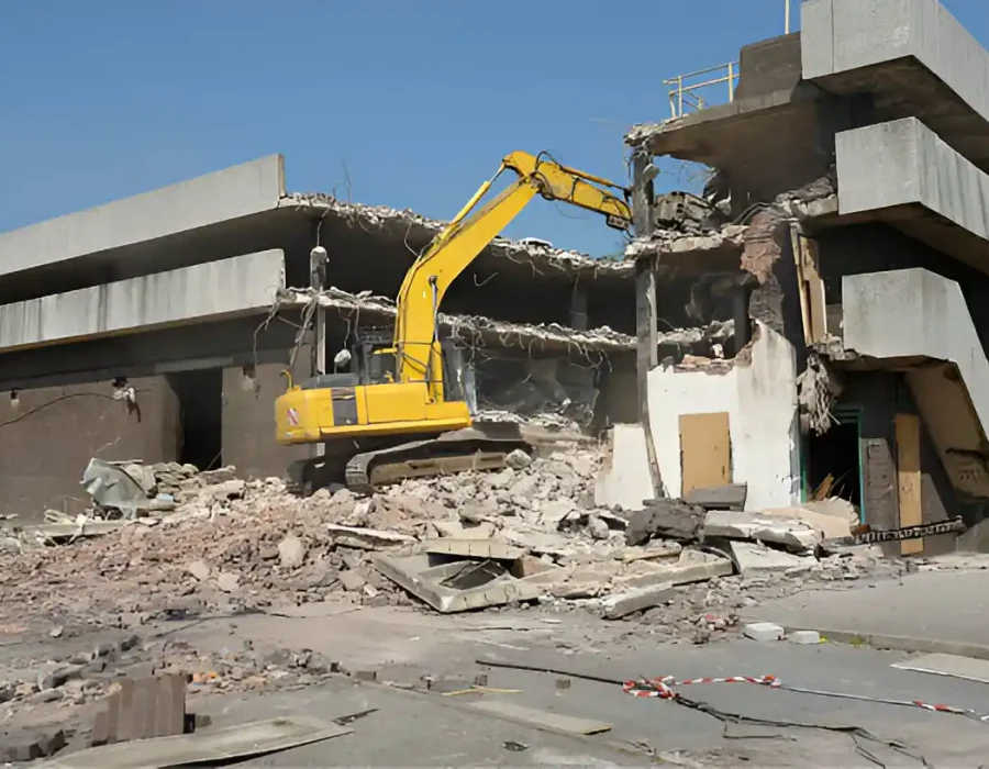 Yellow excavator dismantling concrete building with debris scattered around site