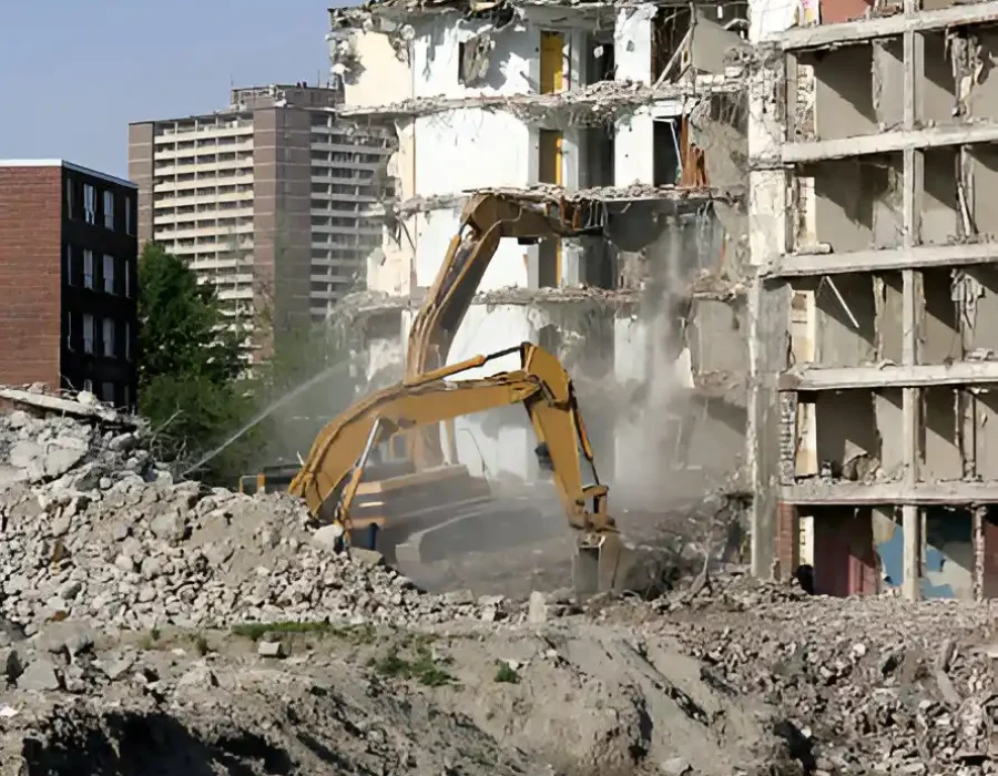 Excavator dismantling multi-story building with debris and surrounding apartments visible