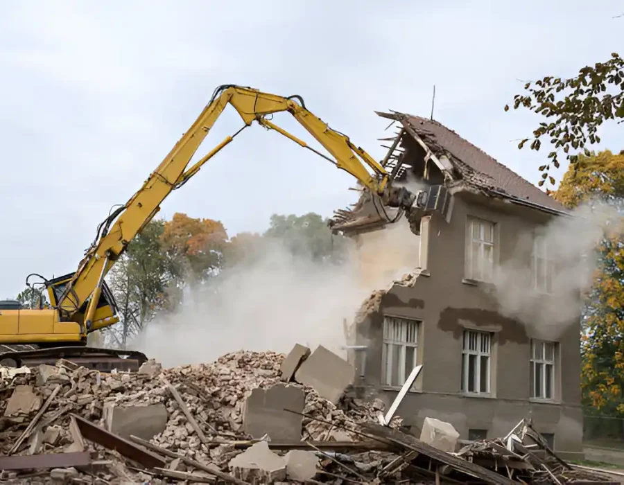 Yellow excavator dismantling two-story house with concrete debris pile
