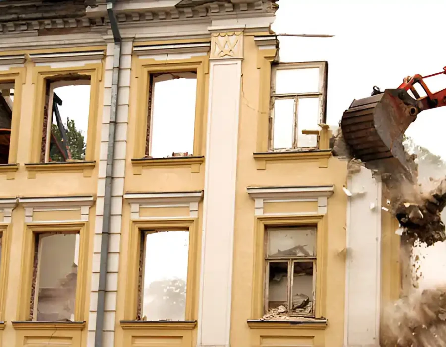 Red excavator dismantling yellow historic building with broken windows